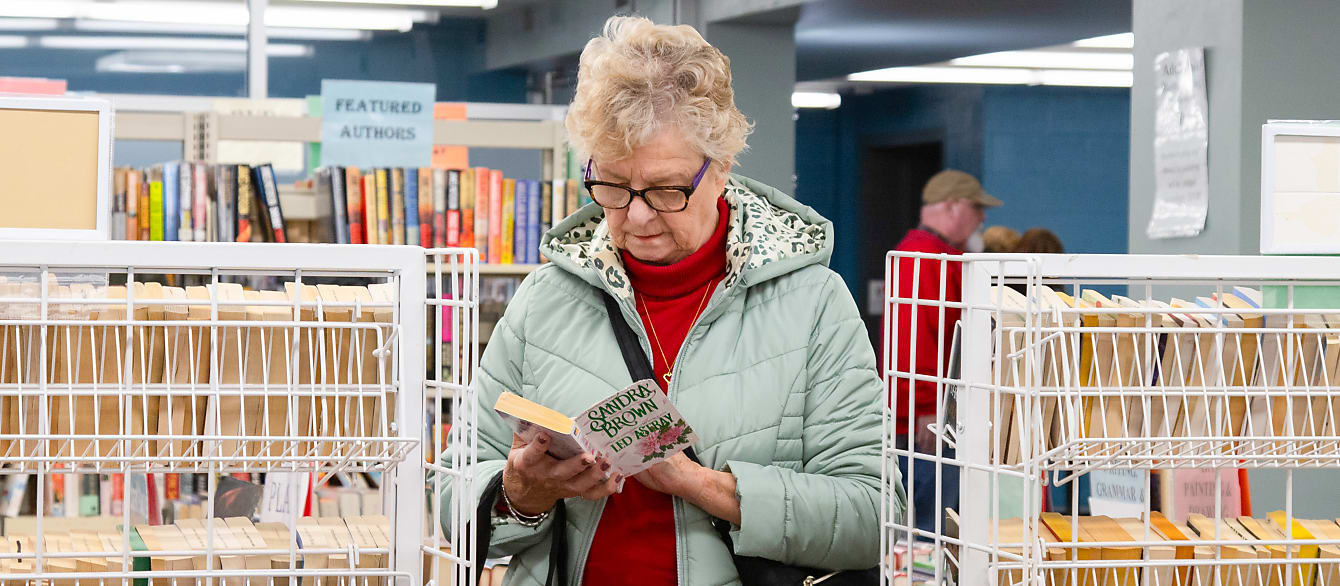A woman in a mint green jacket and red sweater stands between two wire bookshelves while looking inside of a book.