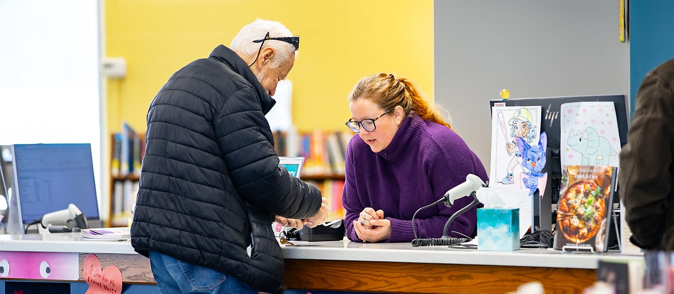 An older man wearing a jacket stands at a help desk and asks for help from the librarian, wearing a purple sweater.