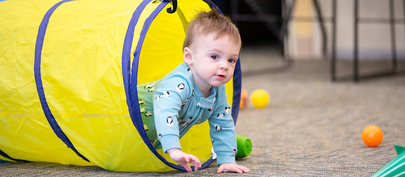 A little boy wearing a penguin onsie crawls out of a yellow play tunnel