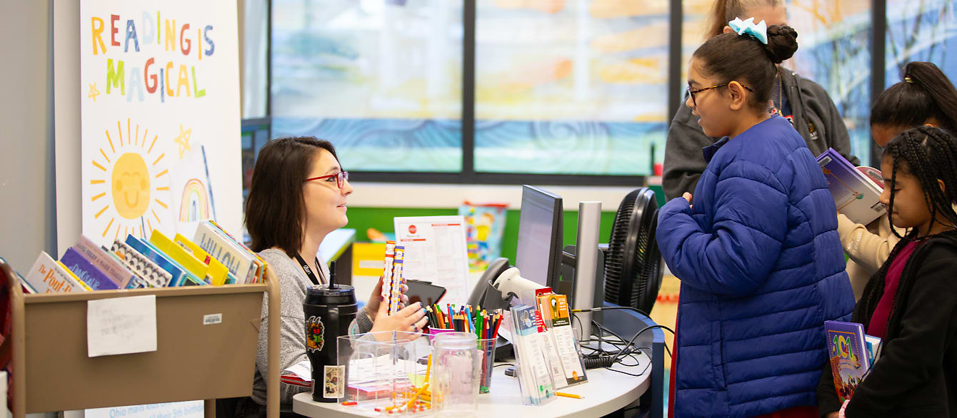 A young girl in a blue puffer jacket stands at the checkout desk with a book in her hands, smiling at the seated librarian.