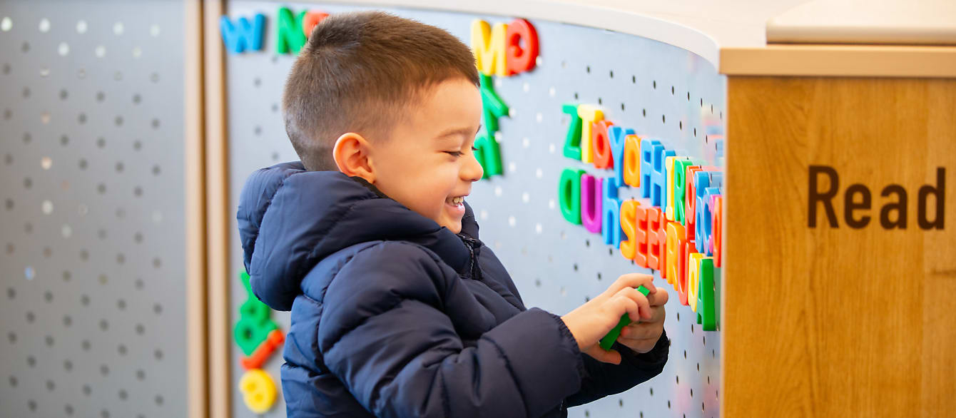 A young boy in a navy blue jacket laughs as he places letter magnets on a metal surface