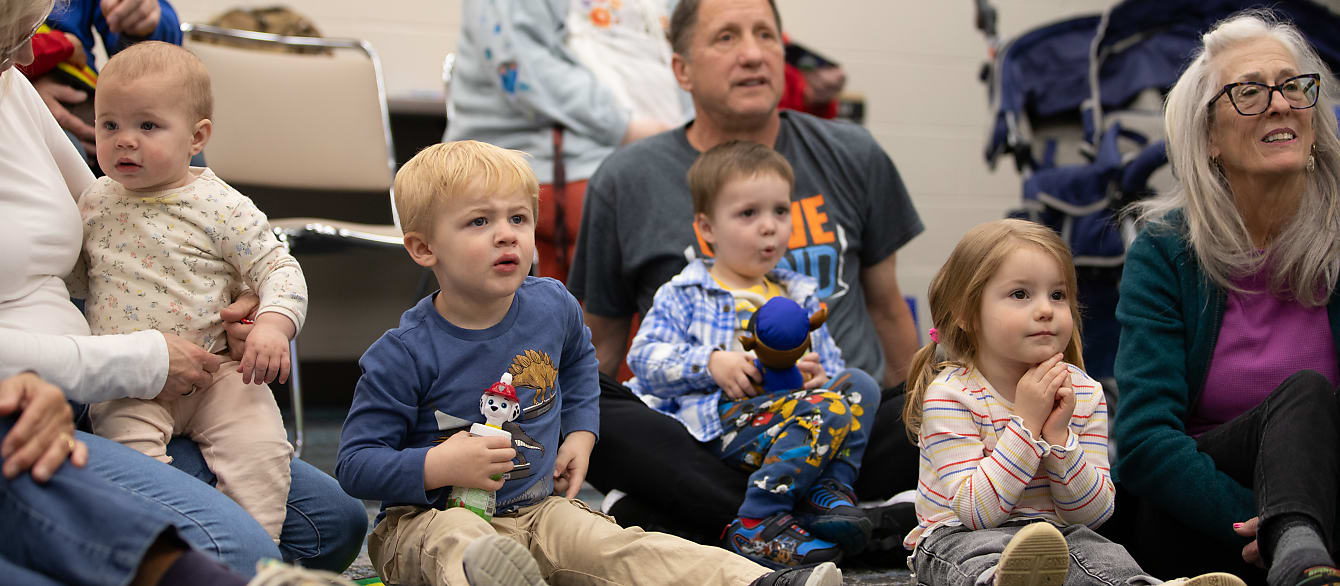 a group of babies, toddlers, and adults listen to a story being read to them.