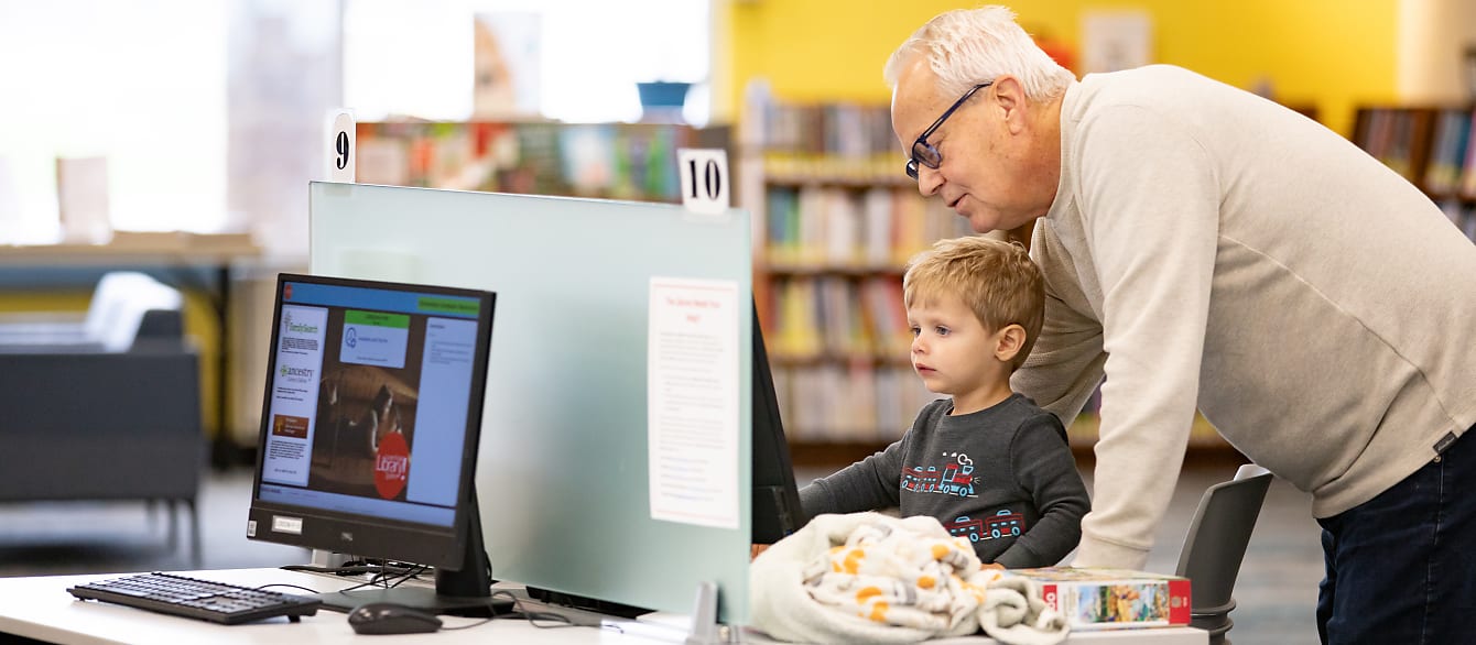 A young boy sits at the computer while his grandfather leans on the table next to him to help him navigate.
