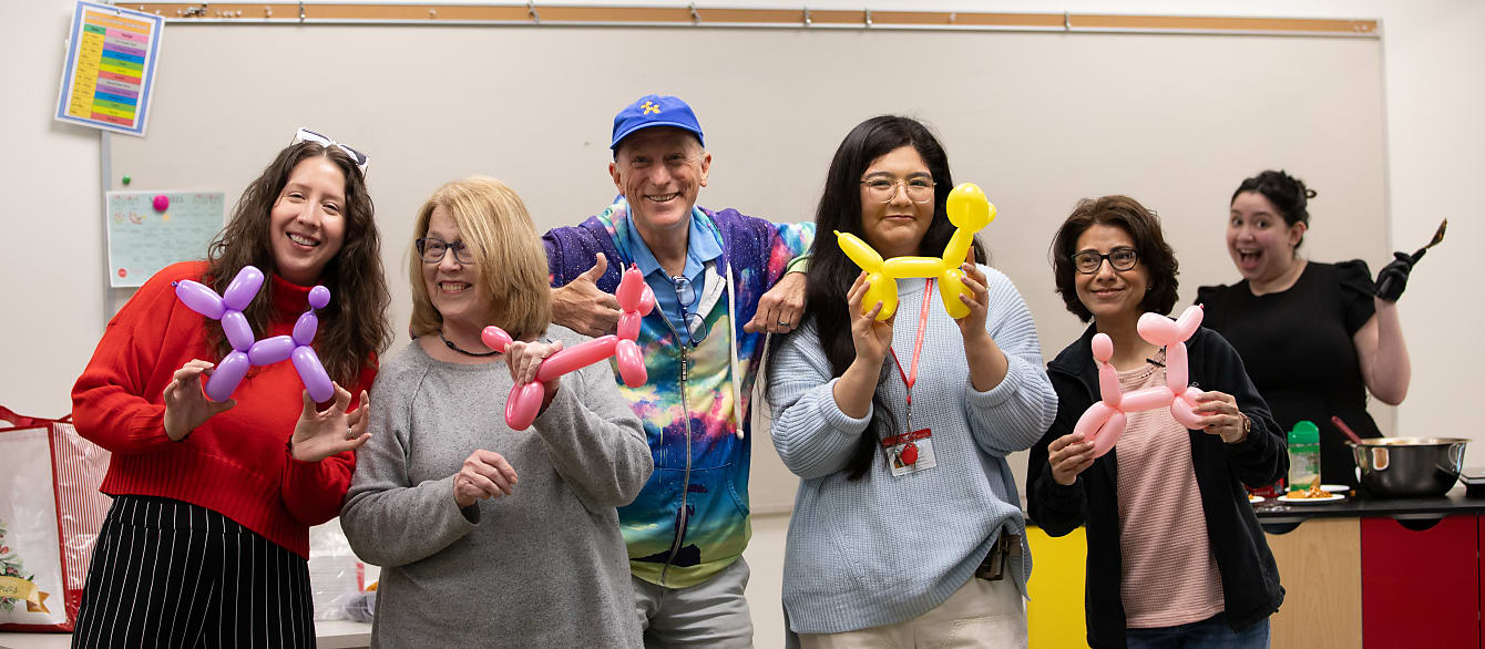 A man in a tie-dye hoodie stands in the middle of a group of women, all holding up balloon dogs and smiling.