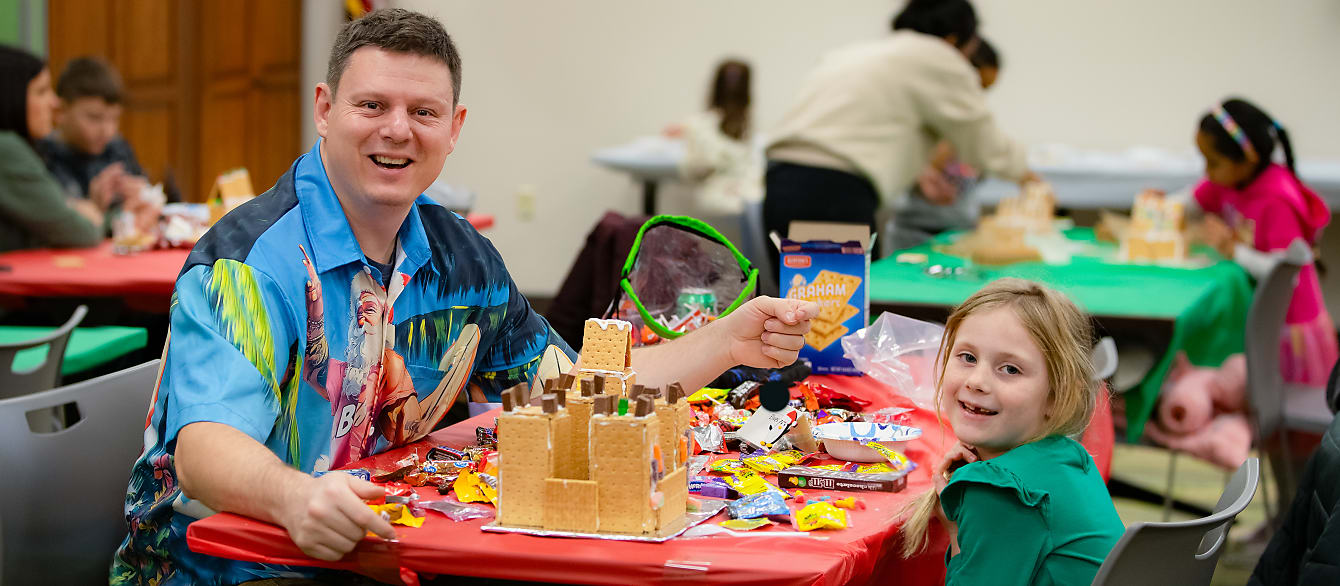 A father and daughter show off thier gingerbread castle while sitting at a table covered with candy 