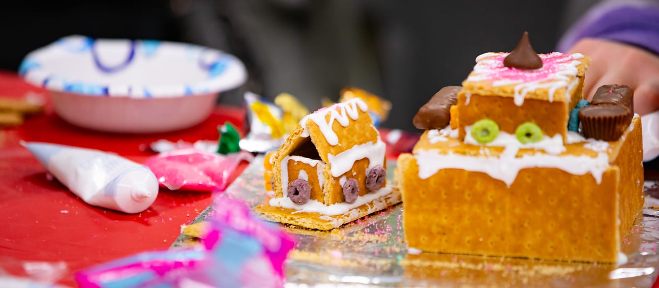A small gingerbread house decorated with frosting and cheerios sits next to a much larger, unfinished house and an icing tube