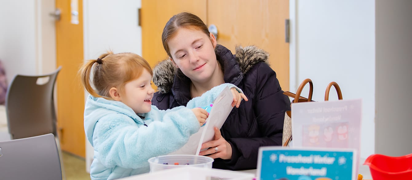 A mom and daughter, both wearing puffer coats, work together to cut out a snowman headband.