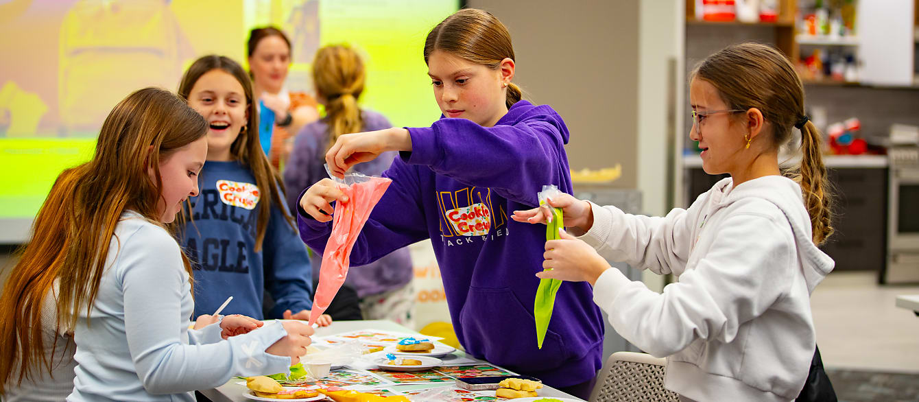 Four tween girls work together to decorate cookies, smiling and holding various piping bags of different colored icing.