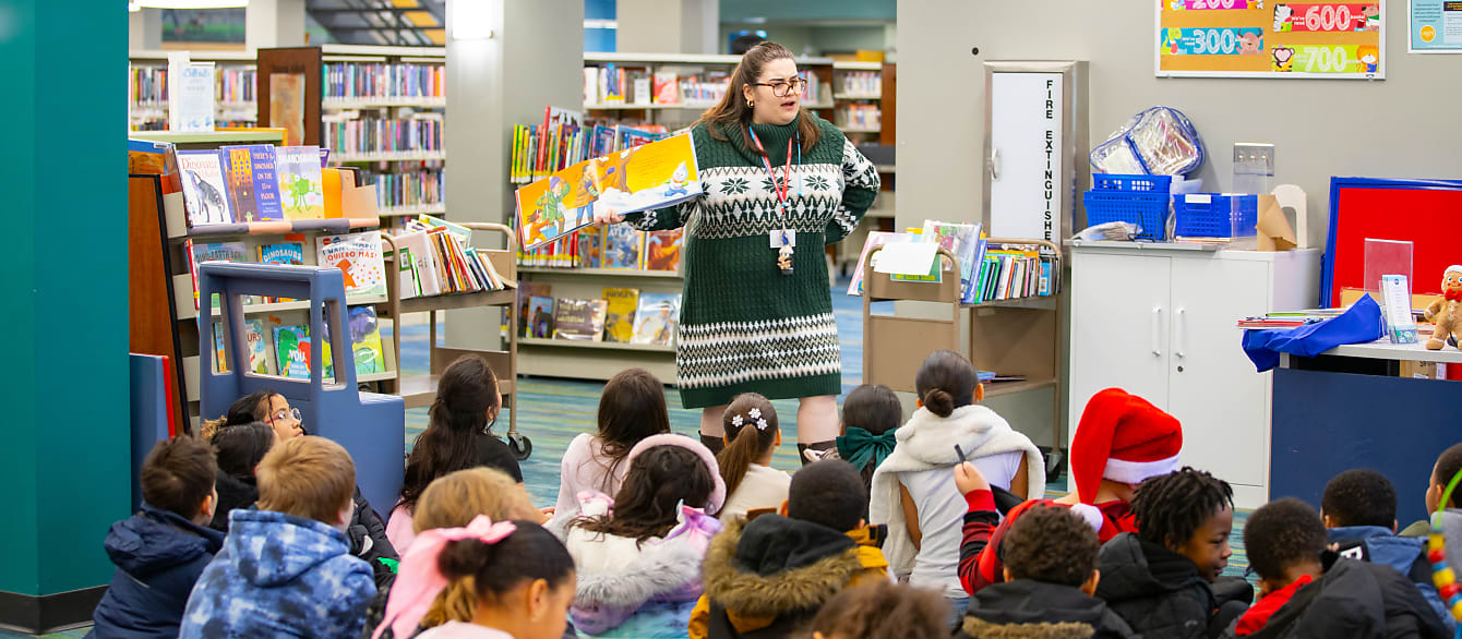 A librarian in a green sweater dress reads to a group of children sitting on the floor in their coats.