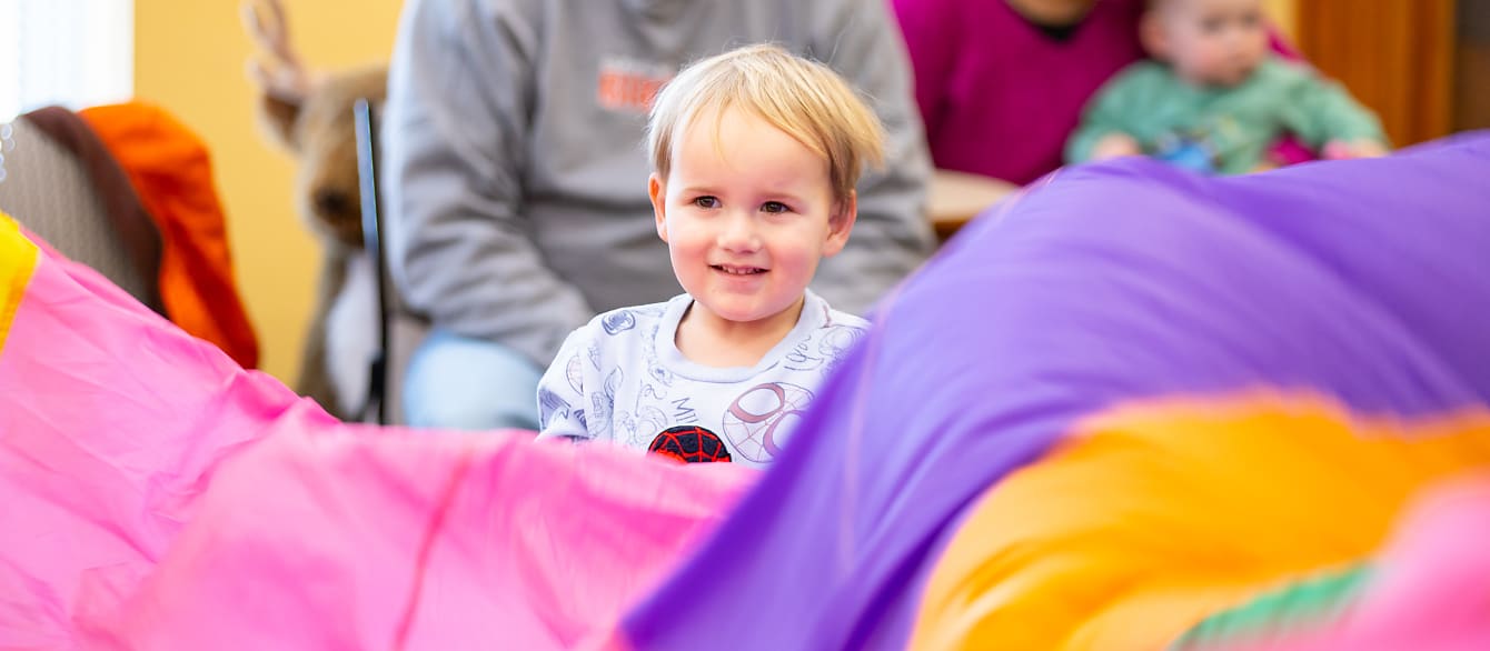 A colorful parachute moves in the foreground while a smiling young boy in a spiderman shirt helps create the motion.