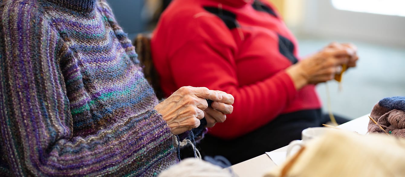 Two women's hands while they are knitting. One in the front wearing a purple sweater and one in the back wearing a red one