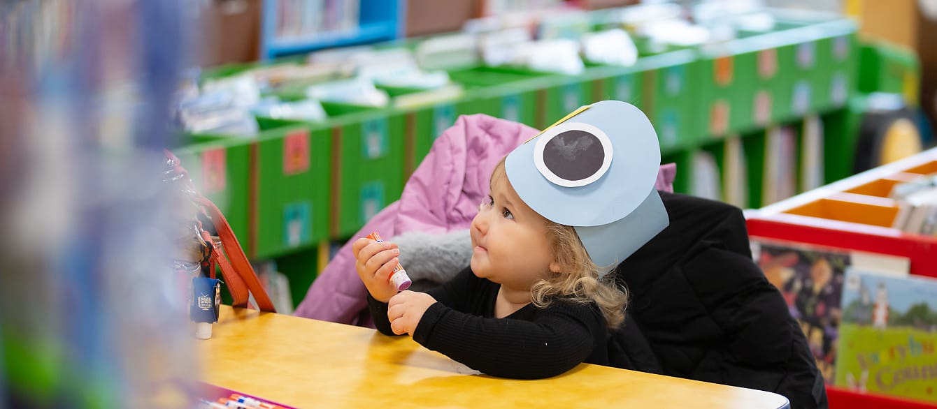 A little girl holding a gluestick looks up while wearing a headband shaped like a Mo Willems pigeon head.