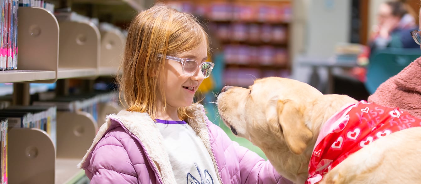 A girl in a pink puffy coat smiles while patting a Labrador Retriever on the head. Their faces are close together.