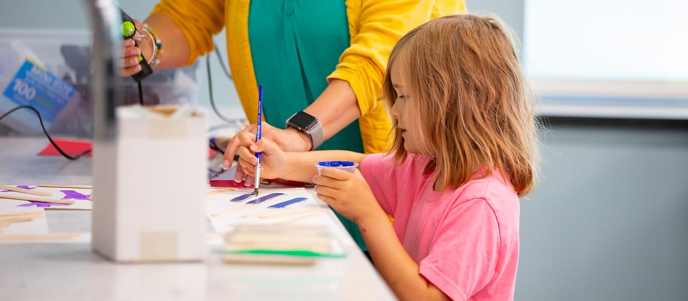 A young girl in a bright pink shirt stands over a counter and paints popsicle sticks dark blue.