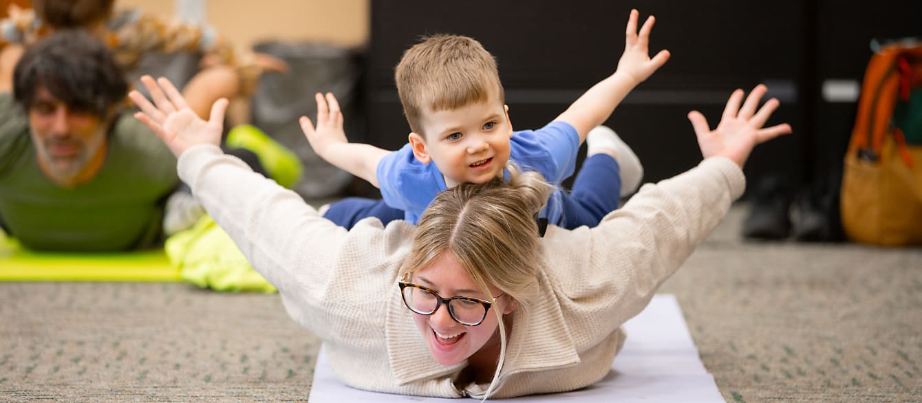 A blonde woman lays on a yoga mat with her arms out with her son laying on her back in the same pose