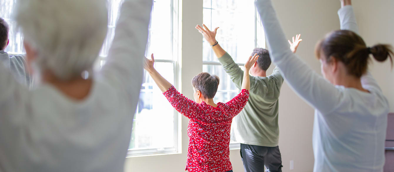 A group of seniors stand in tree pose in front of a wall of windows.