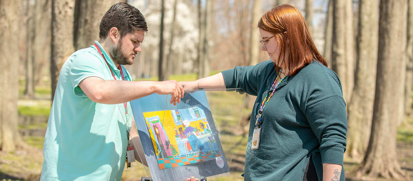 A young man and woman work together to change a panel on a story walk sign outside.