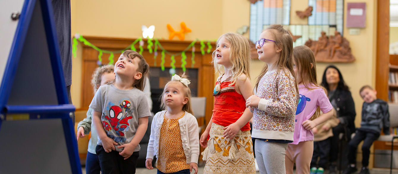 A group of five children, four girls and one boy, ranging from 2-10, gaze up excitedly at the book being read to them. 