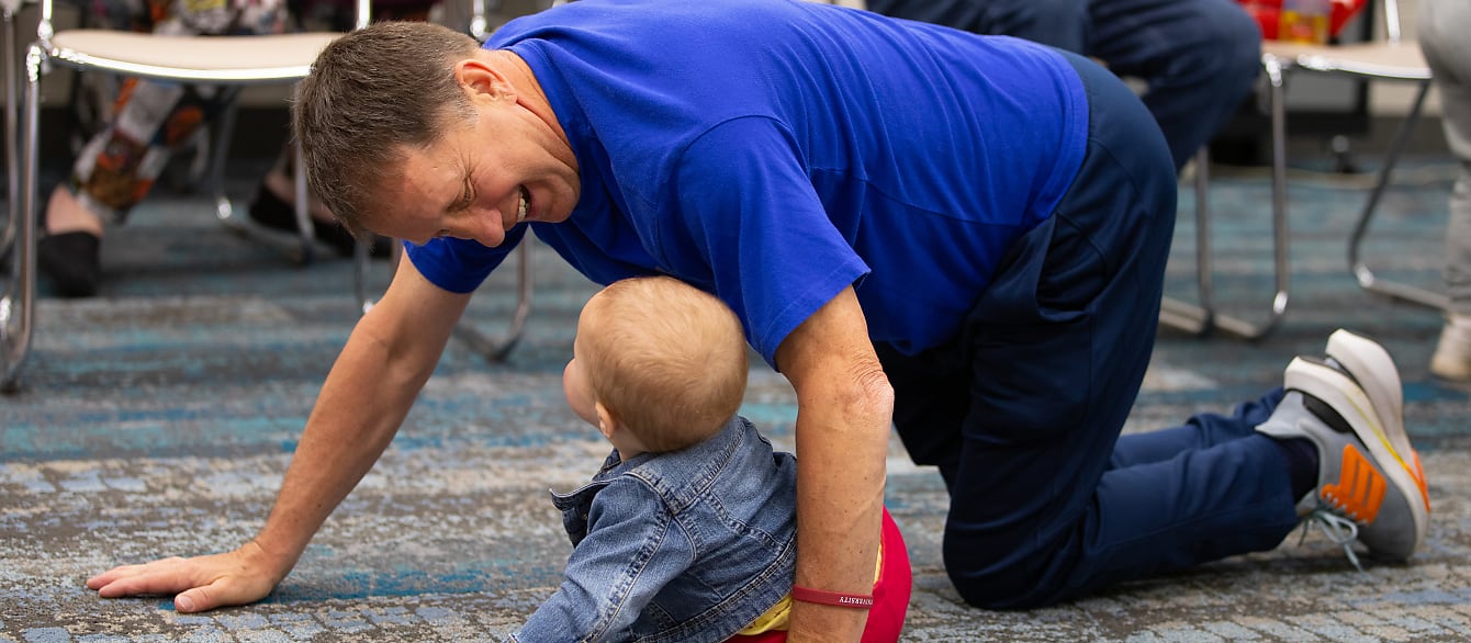 A man in a blue t-shirt crawls alongside a baby in a jean jacket and smiles widely at her.