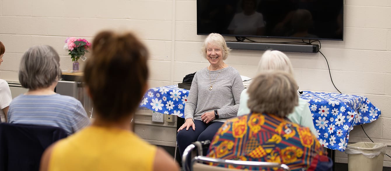 A yoga instructor in a gray sweater smiles at the class of seniors in front of her.