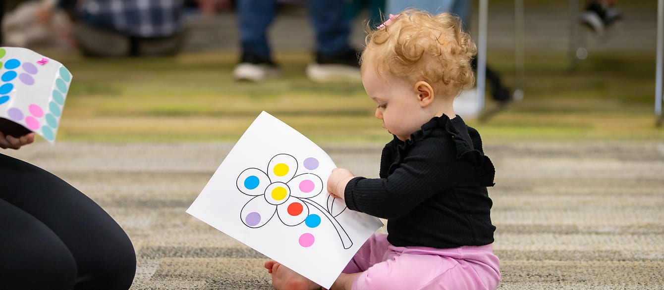 A baby in pink pants and a black shirt places round stickers on a coloring sheet of a flower.