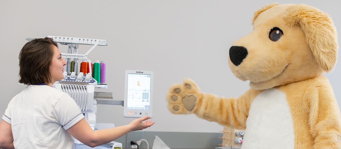 A librarian with short brown hair demonstrates how to use the Embroidery Machine to Browser, the library's dog mascot.