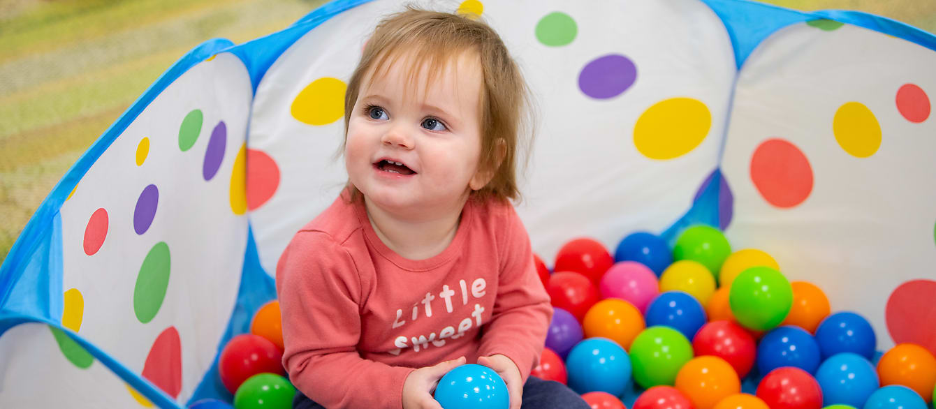 A baby in a pink shirt sits in a small, colorful ball pit, holding a blue ball and smiling at someone off camera.