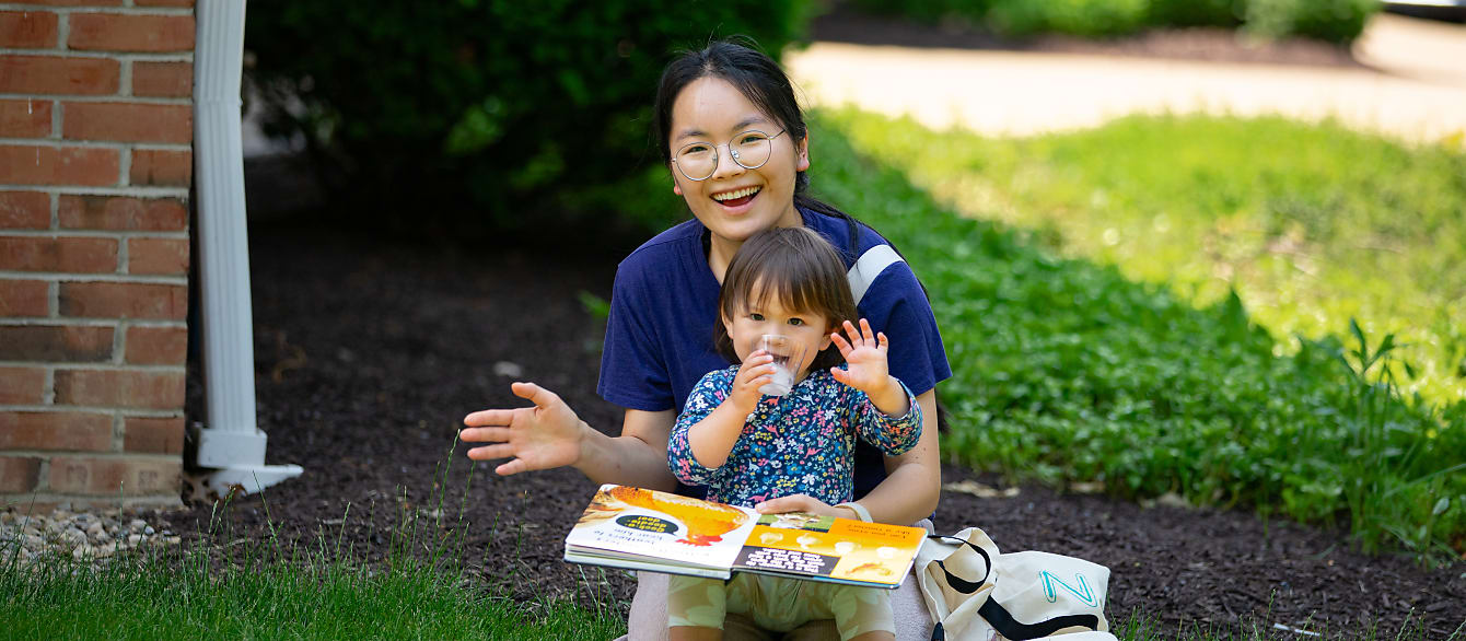 A toddler sits in her mother's lap with an open book, both smiling and waving at the camera.