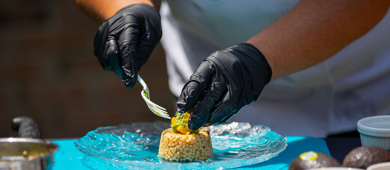 A pair of gloved hands gently place avocado and oranges on top of a small quinoa cake.