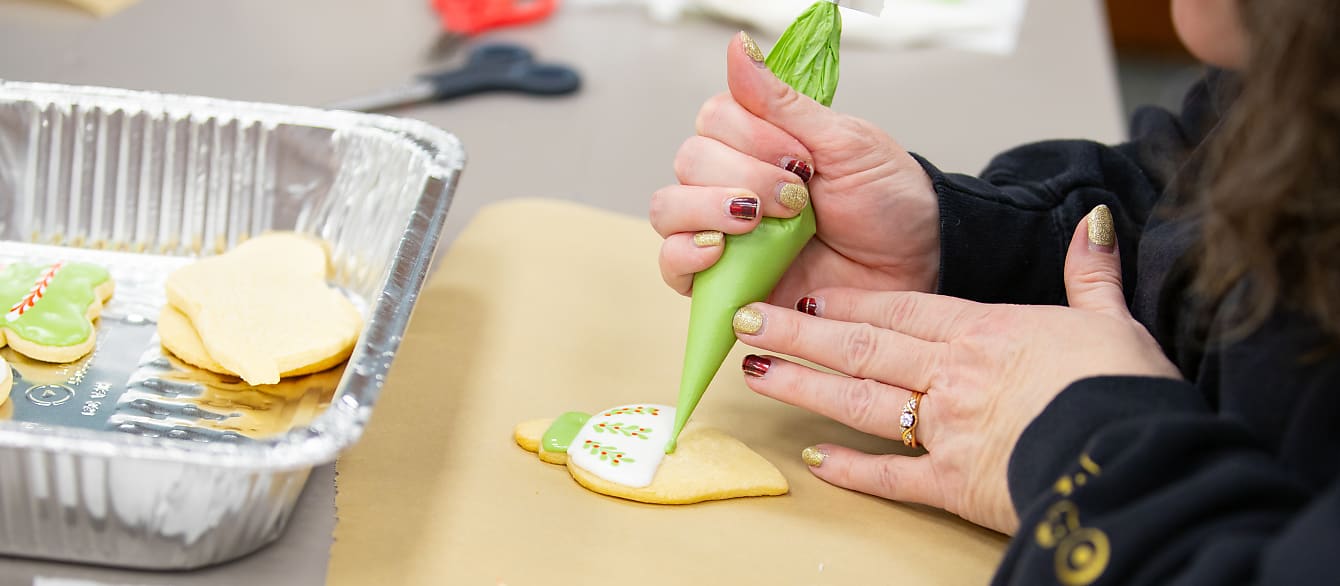 Close-up of a woman's hands with festive nails squeezing green icing onto an ornament-shaped cookie