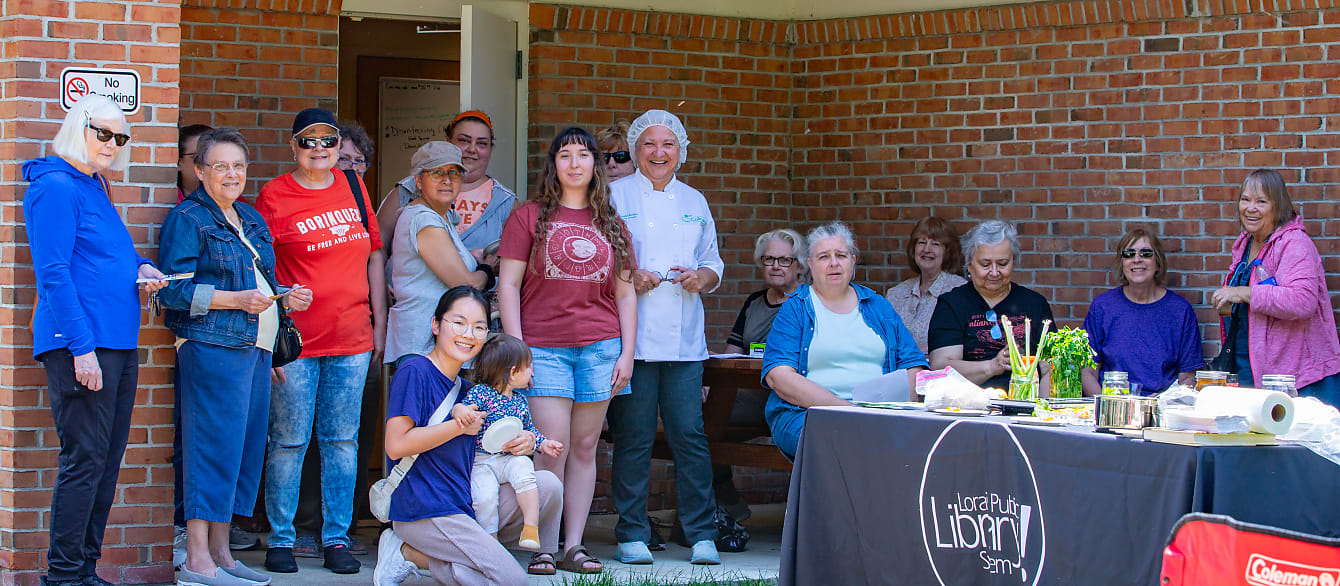 A large group of people stands around a small, portable kitchen outside and smile at the camera.