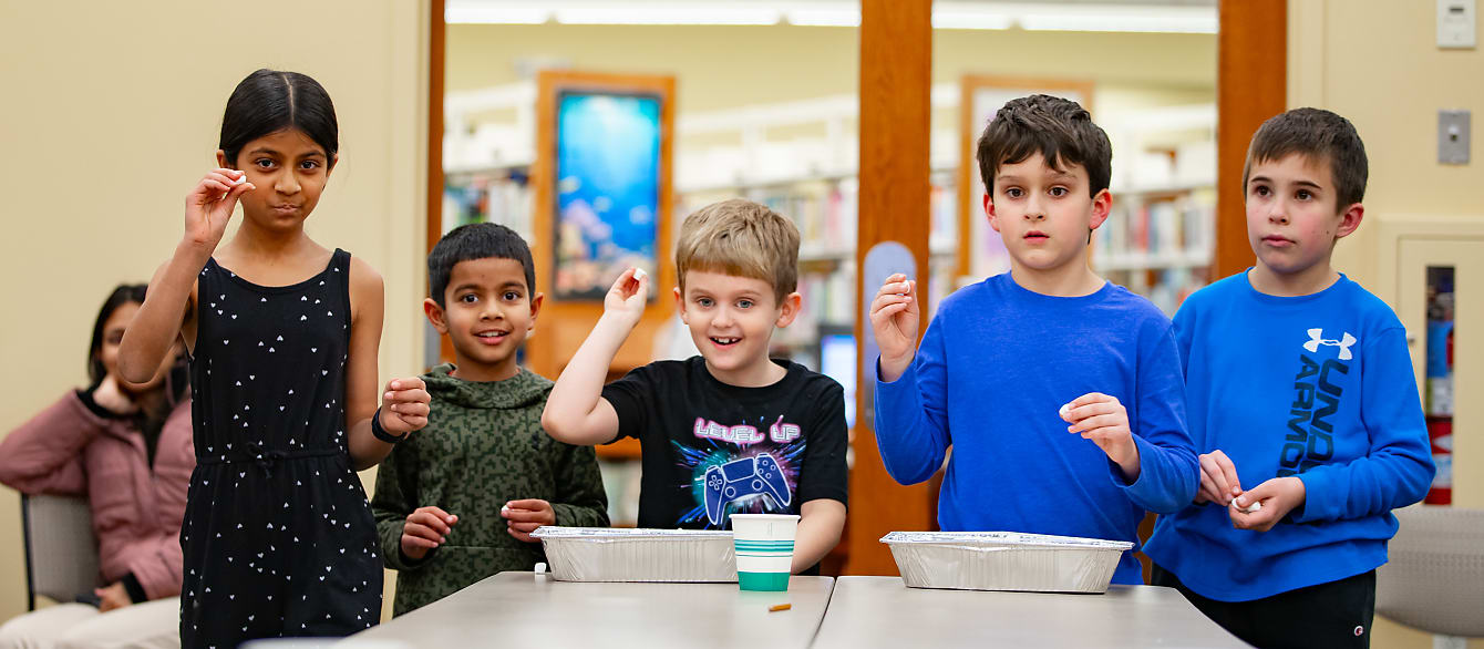A group of five school-age children stand poised with many marshmallows in their hands, waiting for the go-ahead to toss them