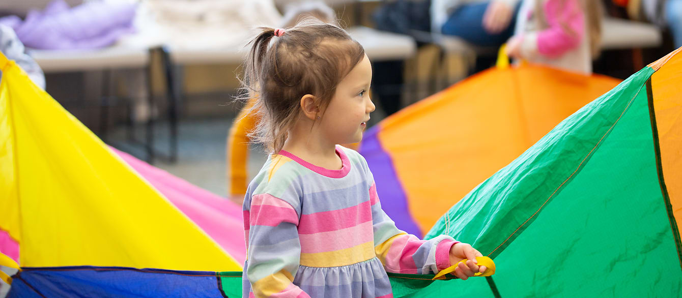 A girl in a striped pink, blue, purple and yellow dress holds on to a colorful play parachute and smiles.