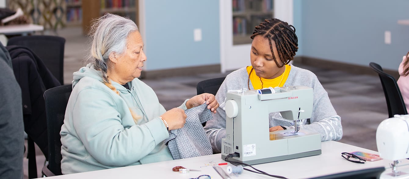 A grandmother demonstrates to her granddaughter where to stitch along the black and white dotted fabric she is holding.