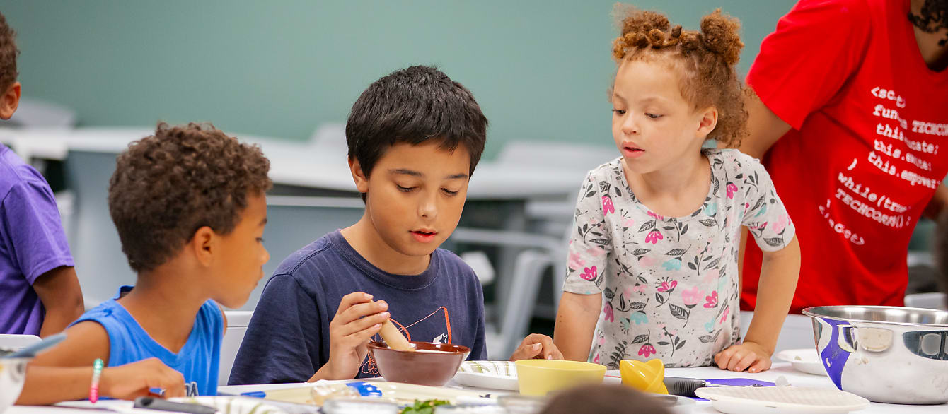Three school-age kids, two boys and one girl, look down at the food they are peeling together