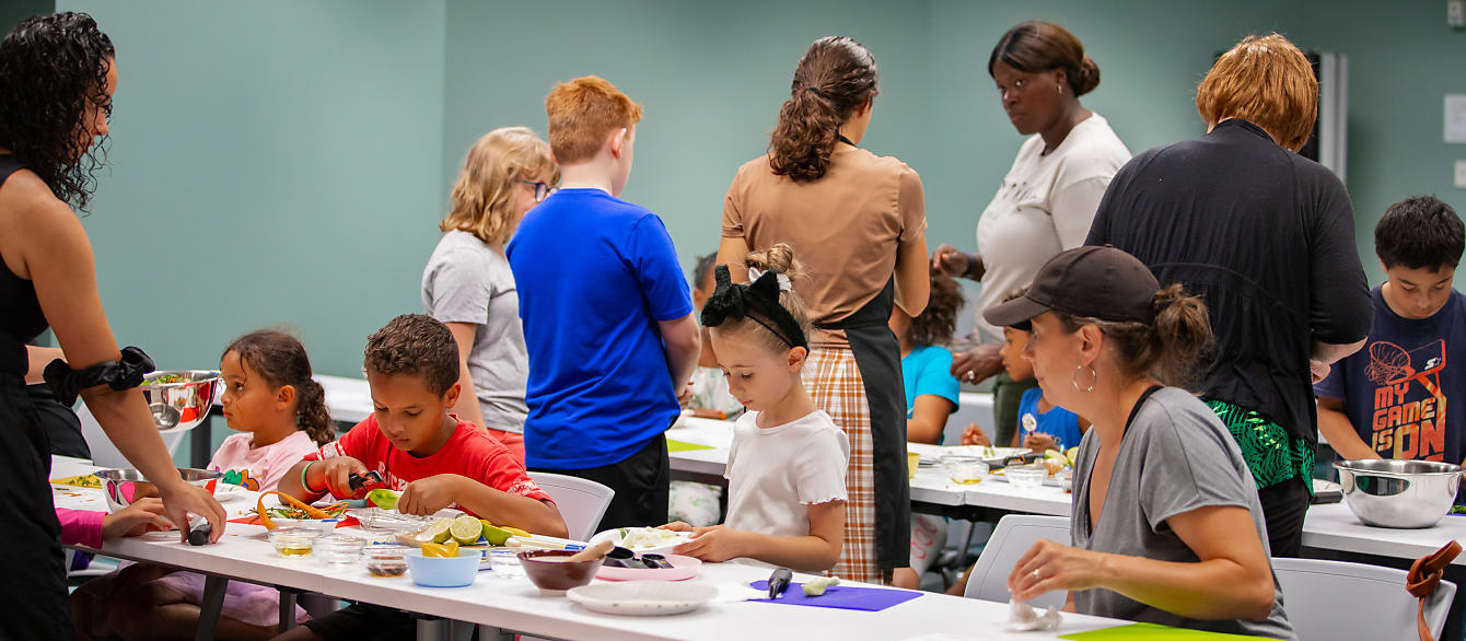 A wide shot of children and caregivers sitting and standing around tables and working with various vegetables.