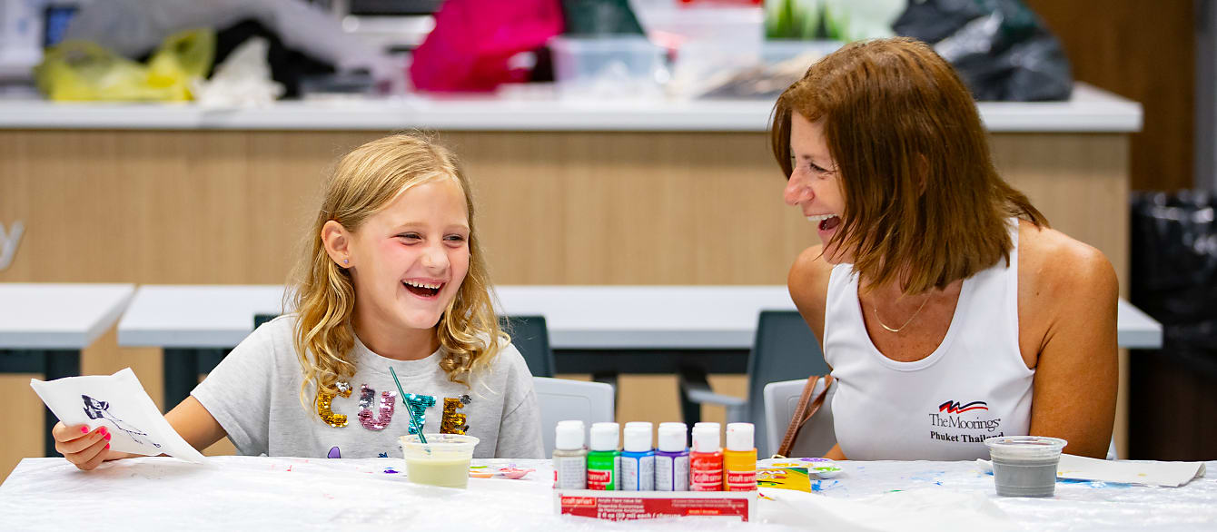 A young girl with blonde hair laughs with her grandmother while sitting at a paint-splattered table.