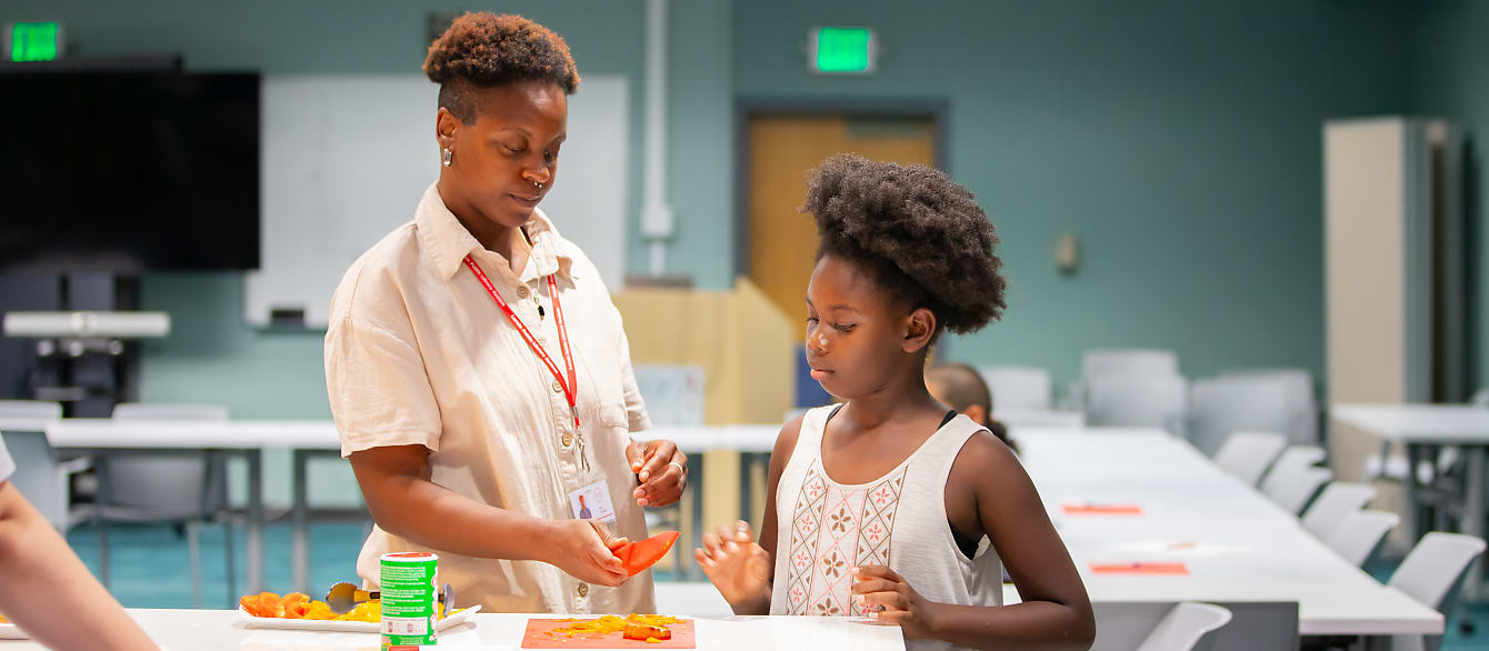 A librarian teaches a young girl how to properly hold a knife to cute sweet peppers.