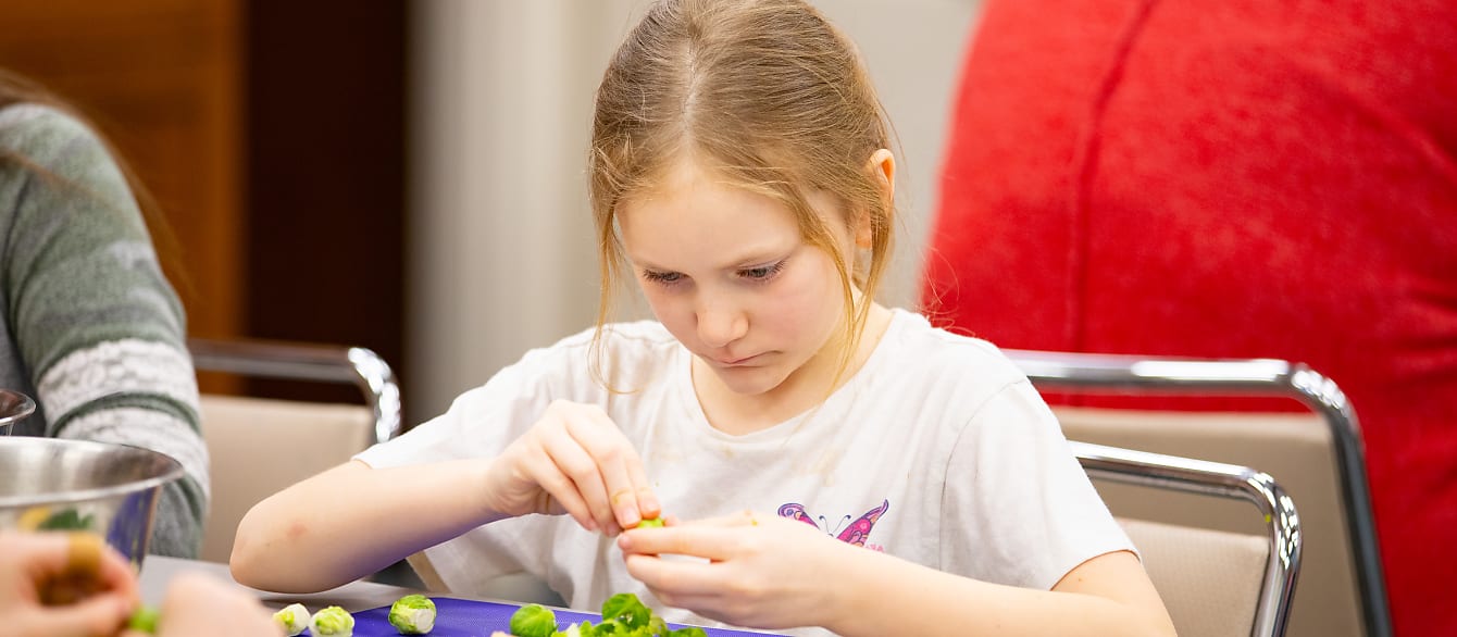 A young girl with blonde hair focuses while peeling a brussel sprout