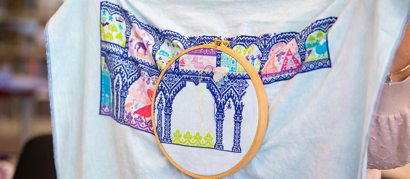 A close-up of someone holding a large, colorful cross stitch project. There is an embroidery hoop in the middle of it.