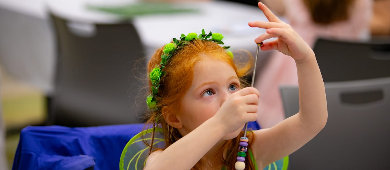 A red-headed little girl wearing a green fair outfit focuses while she loops a bead on a small metal rod.