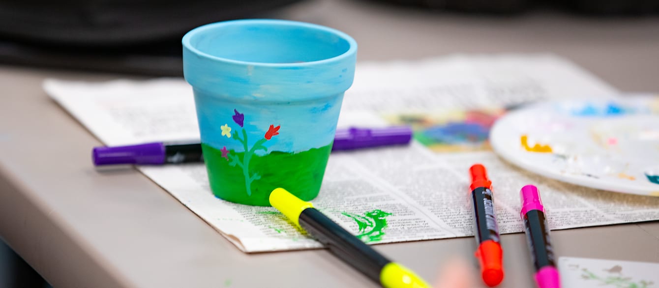 A close-up of a painted flower pot on a newspaper surrounded by acrylic paint markers and paint splotches.