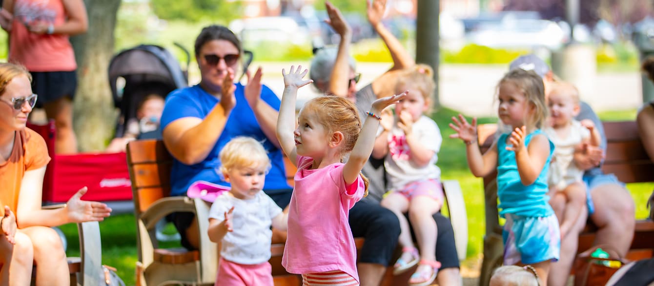 A preschool-age girl with red hair puts her arms up. There is a crowd of people blurry behind her.
