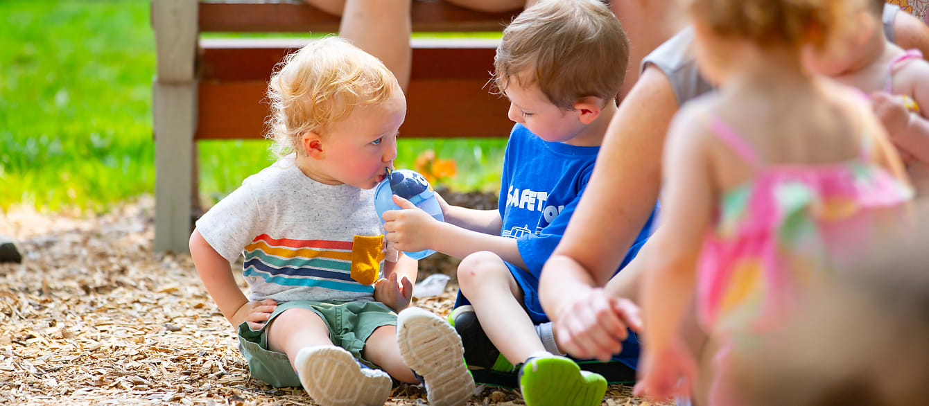 A young boy in a blue shirt offers his baby brother some water from his sippy cup.