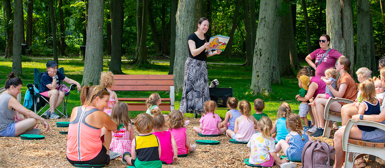 A wide shot of a librarian reading a picture book to a group of children and adults outside.