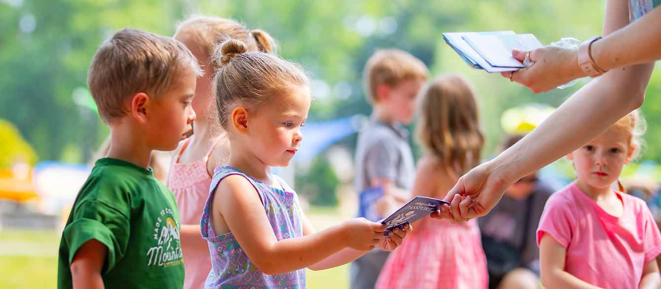 A young girl in a floral tank top receives a story trail passport from an out-of-frame adult.