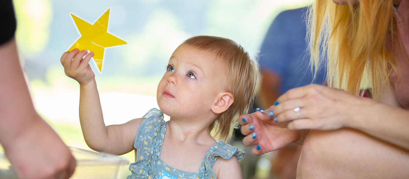 A baby girl holds up a laminated yellow star to show an out-of-frame adult.