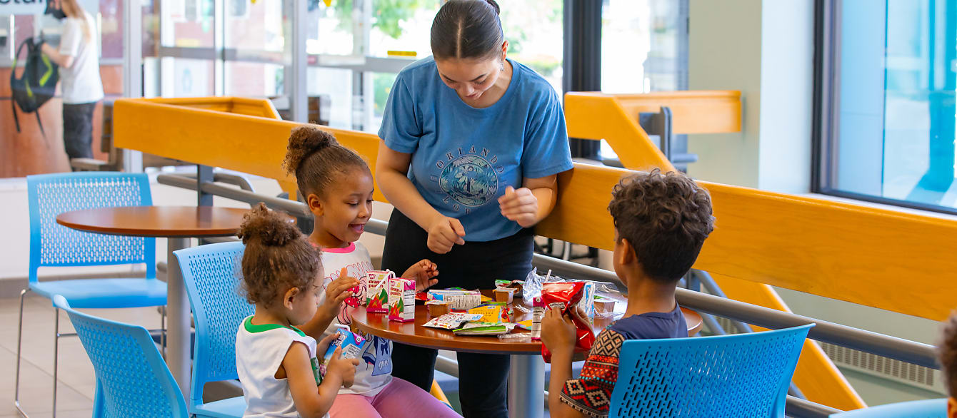Three children sit around a table while an adult doles out lunch.