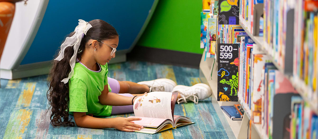 A school-age girl in a neon green shirt lays on the floor reading a book.