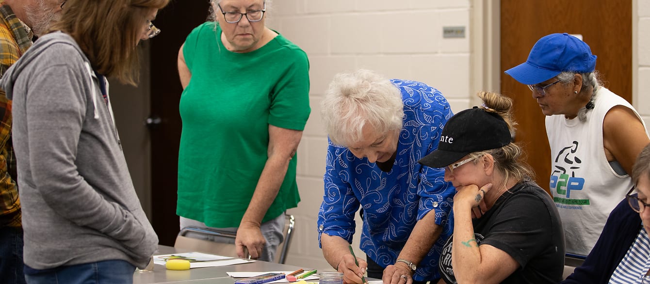 A group of adults stand around an art instructor to learn how to use watercolor pencils.